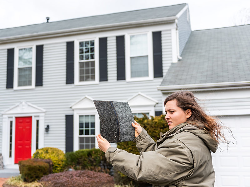 Young female homeowner standing in front of house on windy day holding roof tile shingle inspecting damage